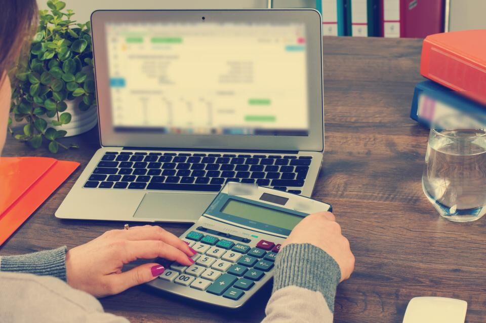 Mãos digitando em uma calculadora ao lado de um notebook com planilha